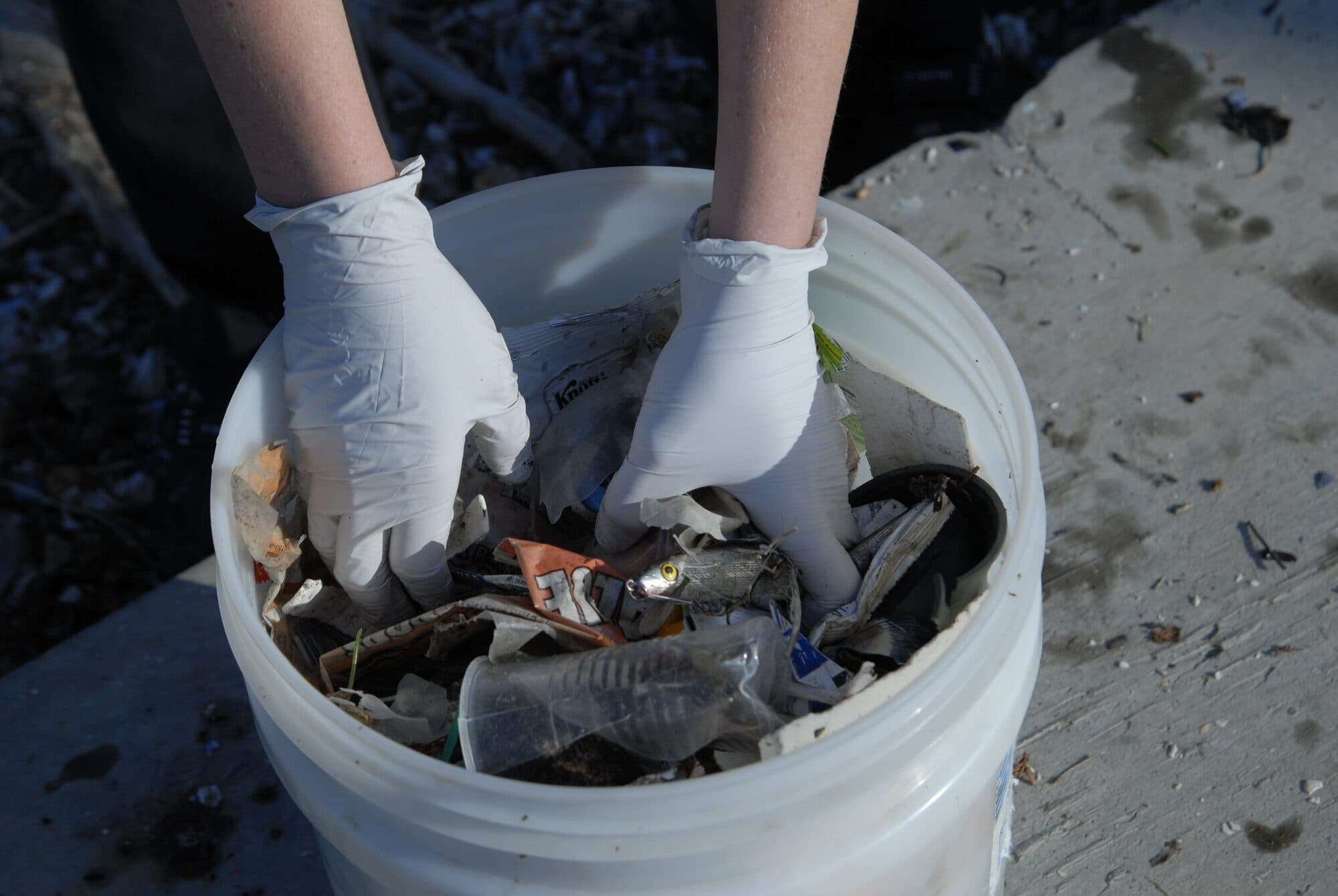 Gloved volunteer placing shoreline trash into a white bucket in Alameda, California