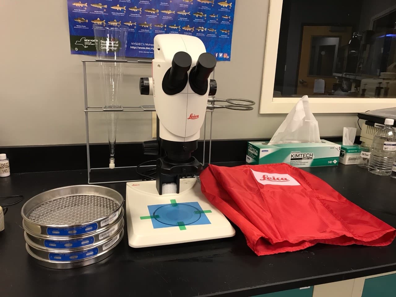 A stereo microscope on a laboratory bench beside a stack of graded sieves and a sample dish